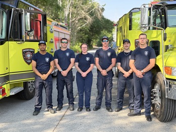 Six firefighters standing between two fire trucks posing for a picture