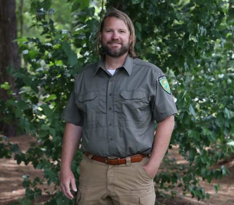 Park Ranger Brent Newman wearing his park ranger uniform and smiling and standing in front of trees in a park.